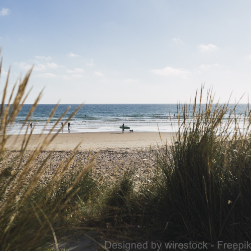 Ein ruhiger Strand mit dem Meer im Hintergrund. Man sieht entfernt einen Surfer am Strand stehen. 