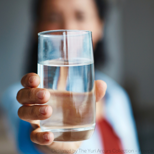 Im Vordergrund ein Glas Wasser, das man scharf sieht. Es wird von einer Frau richtung Kamera gehalten, die man nur unscharf erkennen kann. 