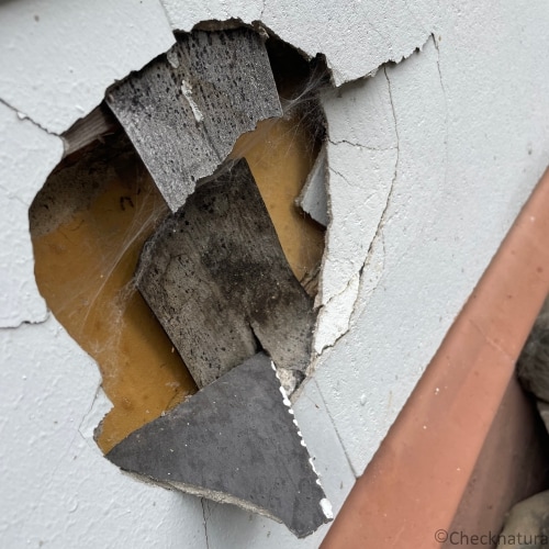 Graue Asbestzementplatten einer Hausfassade mit einem Loch und Bruchkante. 