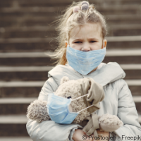 Ein kleines Mädchen trägt eine medizinische Maske und guckt besorgt. Auf dem Arm hat Sie einen Teddy, der ebenfalls eine Atemmaske trägt. 