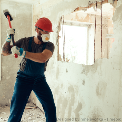 Ein Bauarbeiter mit Atemschutz schlägt mit einem Hammer auf eine Wand, in der sich asbesthaltiges Material befindet.