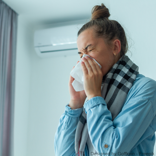 Eine Frau steht vor einer Klimaanlage und niest in ein Taschentuch