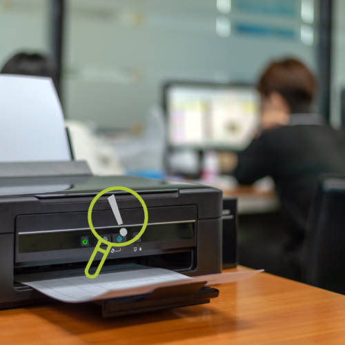 Ein Drucker in einem Büro. Im Hintergrund eine Frau vor einem PC