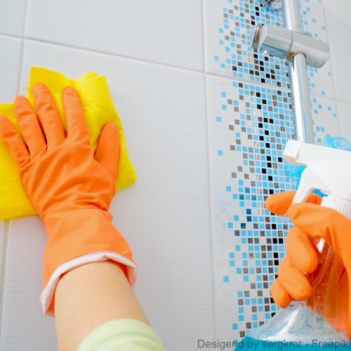 Zwei orangene Handschuhe entfernen Schimmel von einer Kachel im Bad mit Lappen und Spray