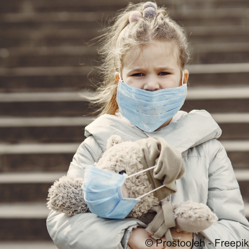 Ein kleines Mädchen trägt eine medizinische Maske und guckt besorgt. Auf dem Arm hat Sie einen Teddy, der ebenfalls eine Atemmaske trägt. 